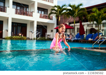 Joyful young girl in pink swimsuit leaps into resort pool. Child enjoys summer vacation swim, playful water jump. Kids active leisure, outdoor family fun. Sunlit holiday villa backdrop. Splash, smile. Joyful young girl in pink swimsuit leaps into resort pool. Child enjoys summer vacation swim, playful water jump. Kids active leisure, outdoor family fun. Sunlit holiday villa backdrop. Splash, smile. 113836582