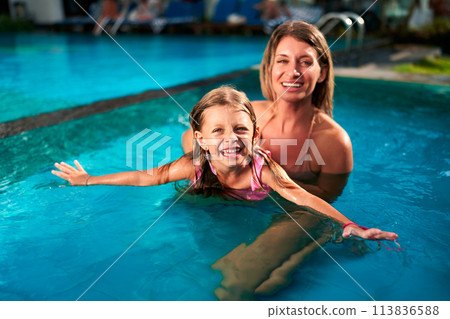 Smiling girl learns swimming in pool with mother support. Child practices paddling, happy mom assists in water safety lesson. Sunny day swim teaching moment, family fun, healthy aqua activity. 113836588