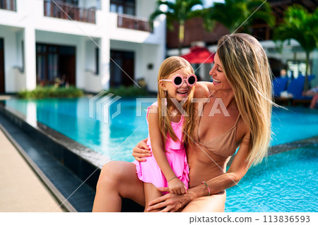 Joyful mother and daughter enjoy sunny poolside moments at luxury resort. Child in pink swimwear, sunglasses smiles with mom in bikini. Family summer holiday fun, lounging by the hotel pool. 113836593
