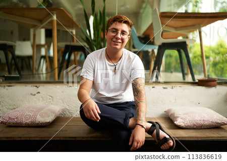 Smiling Gen Z transgender office worker in smart casual clothes sits on bench, modern workplace environment. Tattooed gender-fluid person displays comfort, inclusivity with airy office space. 113836619