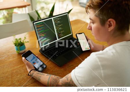 Person at wooden table multitasks with coding on laptop, analyzes data on two smartphones. Concentrated individual with tattoos uses tech for productivity, managing tasks in casual setting. 113836621