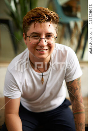 Positive Gen Z transgender person smiles in coworking space. Confident diverse female to male with glasses, tattoos at work. Young adult in casual wear, identity pride at modern office. 113836638