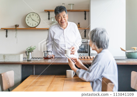 一位老年男子在廚房煮咖啡,一位老年婦女在餐桌上使用智慧型手機 一位老年男子在廚房煮咖啡,一位老年婦女在餐桌上使用智慧型手機 113841109