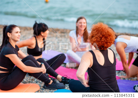 Yoga class. Group of young-adult multi-racial women are sitting on sports mats on wild beach and talking to each other. Concept of female circle of communication and support 113845331