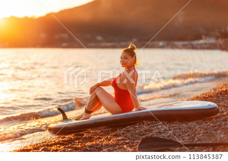 Sexy woman in red swimsuit sitting on sup board at the beach. Model of plus size. The concept of Surfing and summertime 113845387
