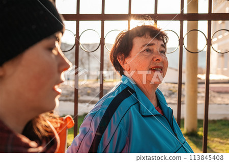 A grandmother and an adult granddaughter are walking down the street. In the background is a fence and. Side view. Family walk in the fresh air A grandmother and an adult granddaughter are walking down the street. In the background is a fence and. Side view. Family walk in the fresh air 113845408