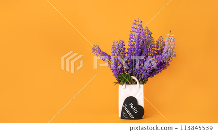 Bouquet of fresh lupine flowers in white vase shaped like bag and hello summer lettering with smile on black heart on orange background 113845539