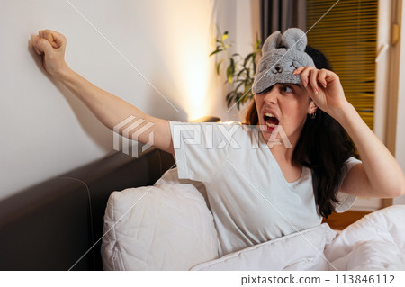 Portrait of annoyance angry Caucasian woman removing sleep mask knocking with fists to wall sitting in bed. Insomnia due to noisy neighbors 113846112