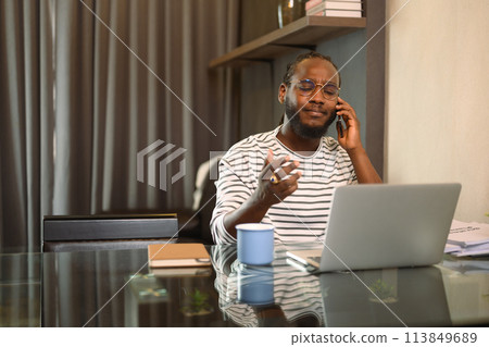Young African man having serious phone conversation and working with laptop at home Young African man having serious phone conversation and working with laptop at home 113849689
