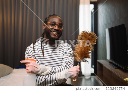 Happy smiling young African guy holding a feather duster ready for cleaning his apartment Happy smiling young African guy holding a feather duster ready for cleaning his apartment 113849754