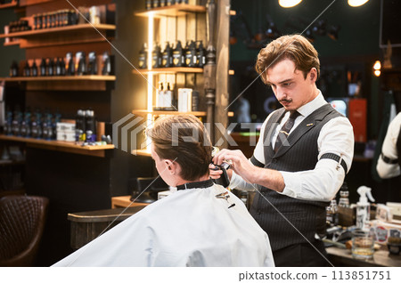 Professional barber using hairspray to finish hairstyle. Stylish hairdresser making final touches. Young customer getting fresh haircut. Professional barber using hairspray to finish hairstyle. Stylish hairdresser making final touches. Young customer getting fresh haircut. 113851751