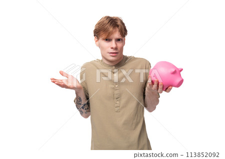 young red-haired man with glasses dressed in a brown shirt holds a pink piggy bank and decides where young red-haired man with glasses dressed in a brown shirt holds a pink piggy bank and decides where 113852092