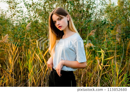 Beautiful young girl wearing blank gray t-shirt and black jeans posing against high green and yellow grass in early warm autumn. Outdoor portrait of beautiful female model. Beautiful young girl wearing blank gray t-shirt and black jeans posing against high green and yellow grass in early warm autumn. Outdoor portrait of beautiful female model. 113852680