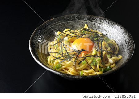 Hot oil soba noodles topped with egg yolk, shredded seaweed, and green onions photographed against a black background 113853051
