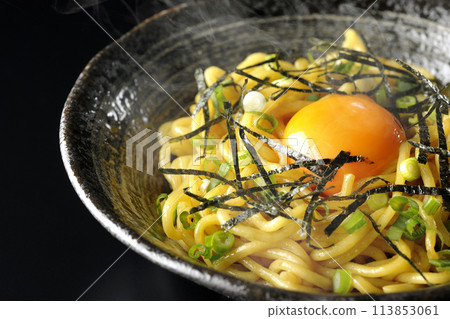 Hot oil soba noodles topped with egg yolk, shredded seaweed, and green onions photographed against a black background 113853061