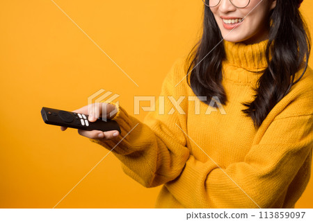 A cozy living room setting with a cheerful young woman in a yellow sweater, happily using a remote control for the television. 113859097