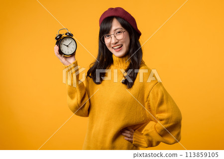 Close up of a vibrant yellow alarm clock and a happy young Asian woman in a fashionable yellow sweater and red beret, exuding a sense of punctuality and elegance against a yellow background. Close up of a vibrant yellow alarm clock and a happy young Asian woman in a fashionable yellow sweater and red beret, exuding a sense of punctuality and elegance against a yellow background. 113859105