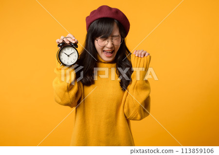A young Asian woman with a happy smile, wearing a yellow sweater and red beret, stands confidently next to an alarm clock in her stylish office. A young Asian woman with a happy smile, wearing a yellow sweater and red beret, stands confidently next to an alarm clock in her stylish office. 113859106