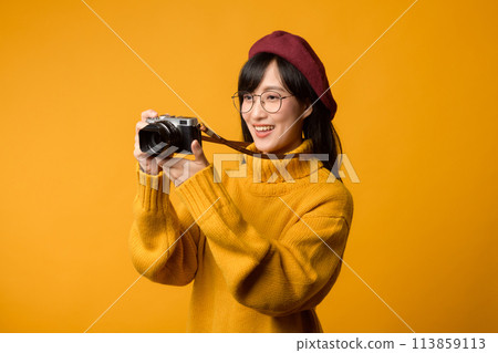 A young Asian woman, a passionate photographer, travels the world with her camera, capturing memories. She wears a yellow sweater and red beret against a vibrant yellow background. 113859113