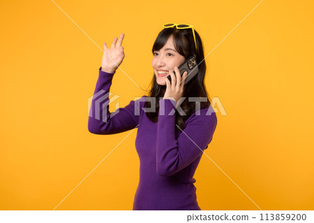 Radiate joy with a young Asian woman in her 30s, wearing an purple shirt, showing okay sign gesture while using smartphone on yellow background. 113859200