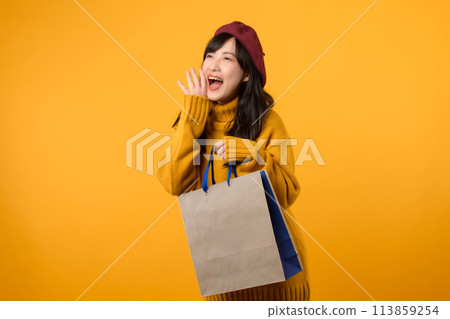 Young Asian woman in her 30s, wearing a yellow sweater and red beret, shouting to free space with shopping paper bag against a vibrant yellow backdrop. Young Asian woman in her 30s, wearing a yellow sweater and red beret, shouting to free space with shopping paper bag against a vibrant yellow backdrop. 113859254