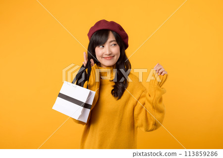 Surprised young Asian woman in her 30s, holding a shopping paper bag, with elegance in a yellow sweater and red beret against a yellow background. 113859286