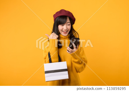 Confident young Asian woman in her 30s, holding a shopping paper bag and smartphone, showcasing her fashionable style in a yellow sweater and red beret against a yellow background, enjoying her 113859295