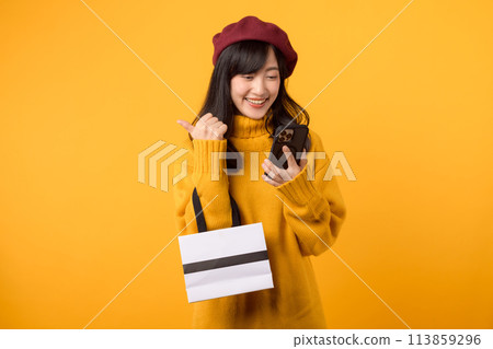 Young Asian woman in her 30s, holding a shopping paper bag and smartphone, celebrating her retail therapy with joy in a yellow sweater and red beret against a yellow background. 113859296
