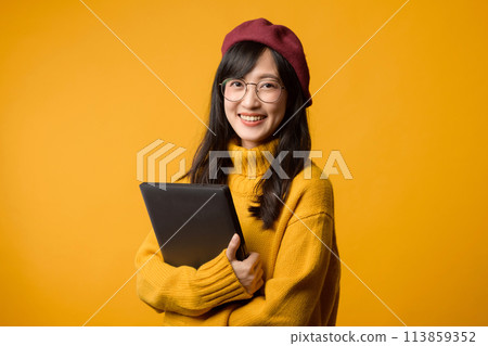 Standing out in a yellow sweater and red beret, a young Asian woman works on her laptop against a cheerful yellow backdrop. 113859352