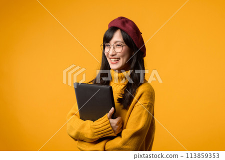 Against a sunny yellow background, a young Asian woman confidently uses her laptop while wearing a yellow sweater and red beret. Against a sunny yellow background, a young Asian woman confidently uses her laptop while wearing a yellow sweater and red beret. 113859353