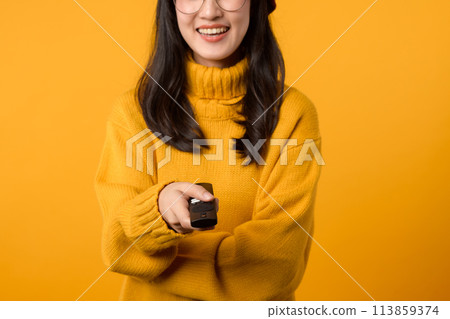 A young Asian woman in a yellow sweater relaxes on her couch with a remote control, indulging in some weekend entertainment. 113859374