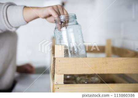 A woman reaches out and places an old glass jar in a recycling bin. A woman collects unwanted glass jars and bottles at home to send for recycling. Glass recycling concept. A woman reaches out and places an old glass jar in a recycling bin. A woman collects unwanted glass jars and bottles at home to send for recycling. Glass recycling concept. 113860386