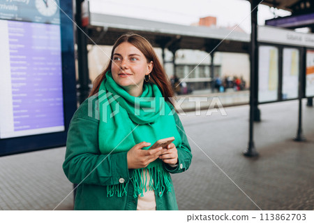 Young redhead woman with backpack holding phone near train timetable on a railway station. 30s female posing on Arrival and Departure Information Display background, Traveler Young redhead woman with backpack holding phone near train timetable on a railway station. 30s female posing on Arrival and Departure Information Display background, Traveler 113862703