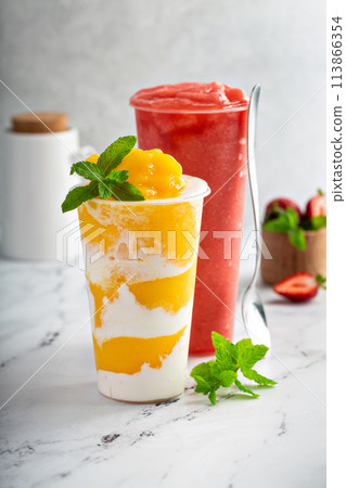 Mango smoothie with whipped coconut cream swirled in a tall cup and strawberry smoothie 113866354