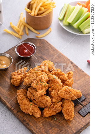 Chicken nuggets or chicken strips on a board with dipping sauces, fries and celery 113867820