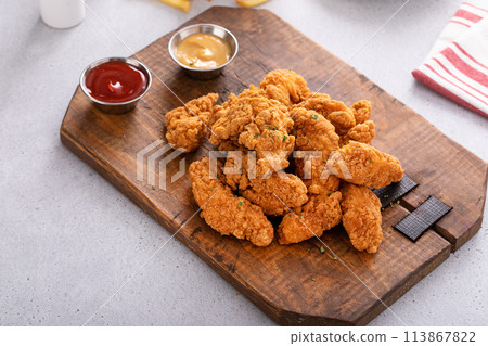 Chicken nuggets or chicken strips on a board with dipping sauces, fries and celery 113867822