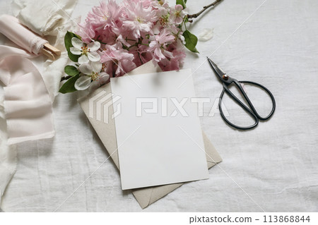 Wedding spring styled photo. Feminine scene mockup with pink blossoming Japanese cherry, apple tree branch. Blank paper greeting card, black scissors, white linen table background. Flat lay, top view. Wedding spring styled photo. Feminine scene mockup with pink blossoming Japanese cherry, apple tree branch. Blank paper greeting card, black scissors, white linen table background. Flat lay, top view. 113868844