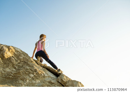 Woman hiker climbing steep big rock on a sunny day. Young female climber overcomes difficult climbing route. Active recreation in nature concept. 113870964