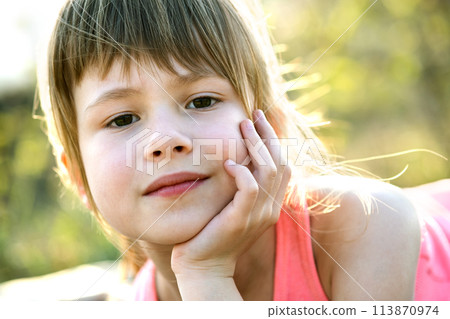 Portrait of pretty child girl with gray eyes and long fair hair leaning on her hands smiling happily outdoors on blurred bright background. Cute female kid on warm summer day outside. Portrait of pretty child girl with gray eyes and long fair hair leaning on her hands smiling happily outdoors on blurred bright background. Cute female kid on warm summer day outside. 113870974