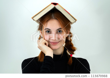 Portrait of funny young smiling student girl with an open book on her head. Reading and education concept. Portrait of funny young smiling student girl with an open book on her head. Reading and education concept. 113870988