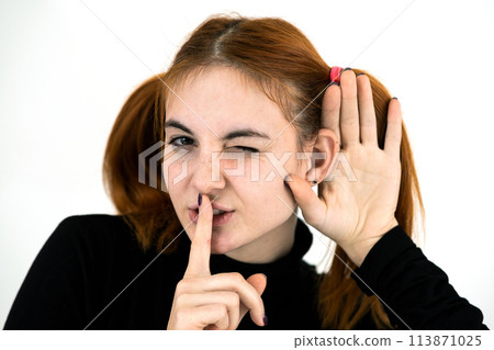 Close up portrait of pretty redhead teenage girl holding a hand to her ear listening to a secret. 113871025