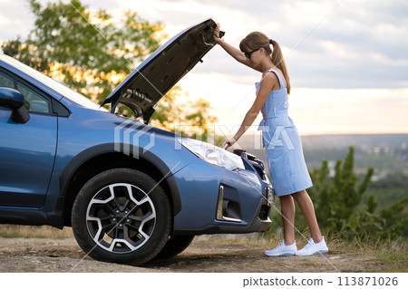 Puzzled young female driver standing near a broken car with open up hood inspecting her vehicle motor. 113871026