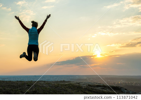 Silhouette of a woman hiker jumping alone on empty field at sunset in mountains. Female tourist raising her hands up in evening nature. Tourism, traveling and healthy lifestyle concept. Silhouette of a woman hiker jumping alone on empty field at sunset in mountains. Female tourist raising her hands up in evening nature. Tourism, traveling and healthy lifestyle concept. 113871042