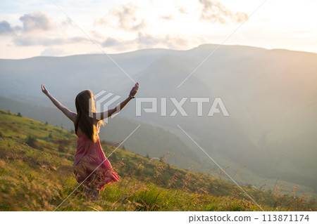 Back view of young happy woman traveler in red dress standing on grassy hillside on a windy evening in summer mountains with outstretched arms enjoying view of nature at sunset. Back view of young happy woman traveler in red dress standing on grassy hillside on a windy evening in summer mountains with outstretched arms enjoying view of nature at sunset. 113871174
