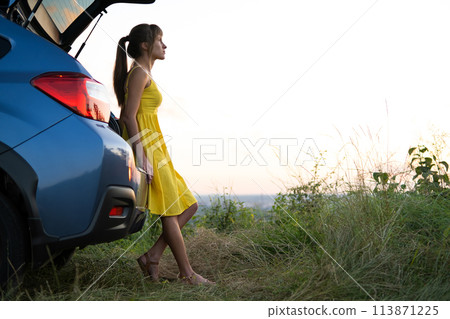 Happy young woman in yellow dress standing near her vehicle looking at sunset view of summer nature. Travelling and vacation concept. 113871225