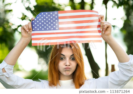 Angry young red haired woman protester posing with USA national flag up over her head standing outdoors in summer park. Activist girl on demonstration with national banner. 113871264
