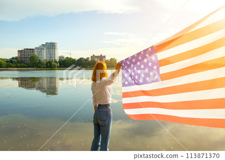 Young womanl with USA national flag on her shoulders with high city buildings in background celebrating United States independence day. 113871370
