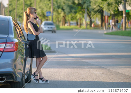 Young female driver standing near her car talking on mobile phone on a city street in summer 113871407