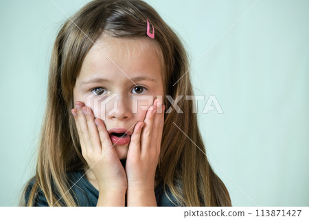 Close-up portrait of little child girl with long hair covering her mouth with hands. 113871427