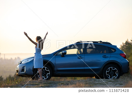 Happy young woman driver in blue dress enjoying warm summer evening standing beside her car. Travelling and vacation concept. Happy young woman driver in blue dress enjoying warm summer evening standing beside her car. Travelling and vacation concept. 113871428